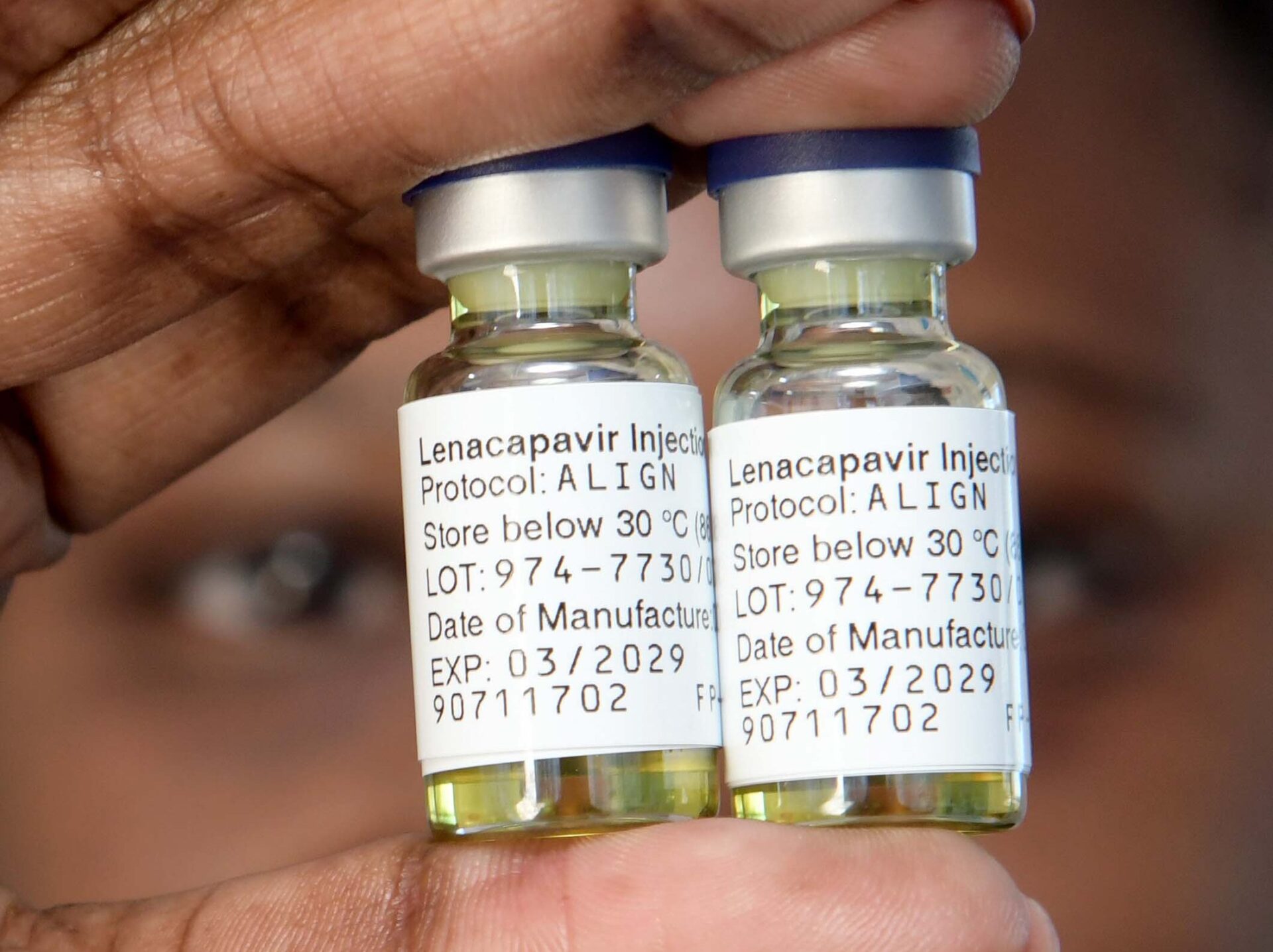 A close-up of a hand, with red, painted fingernails, holding two vials of the lenacapavir injection.